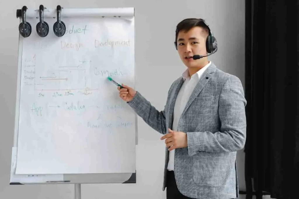 Formateur en blazer expliquant un schéma sur tableau : une session de formation closer high ticket axée sur stratégie, produit et développement.