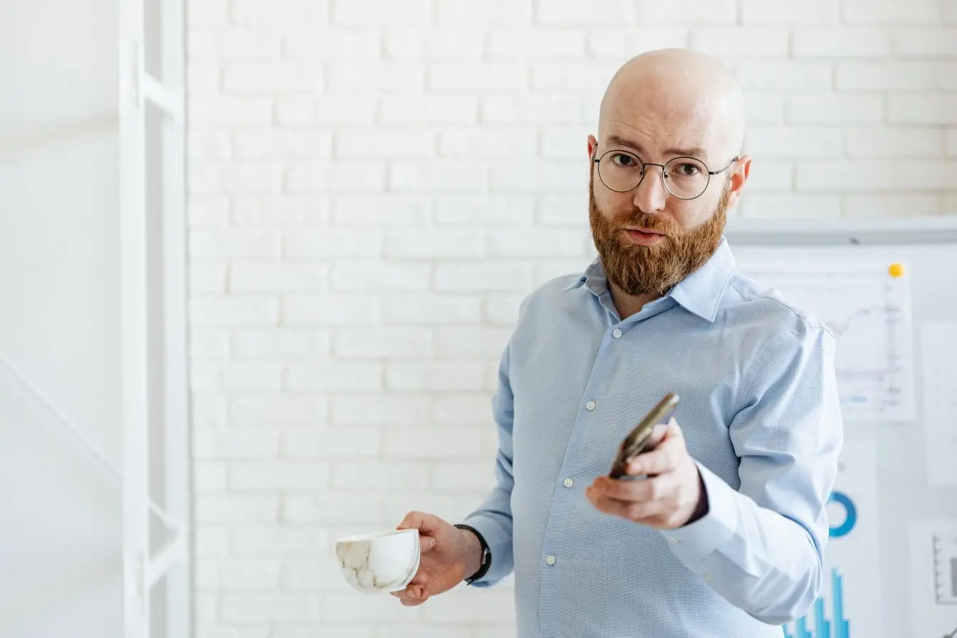 Homme en chemise bleue tenant smartphone et tasse devant graphiques, illustrant une communication volontaire et ciblée basée sur le consentement.
