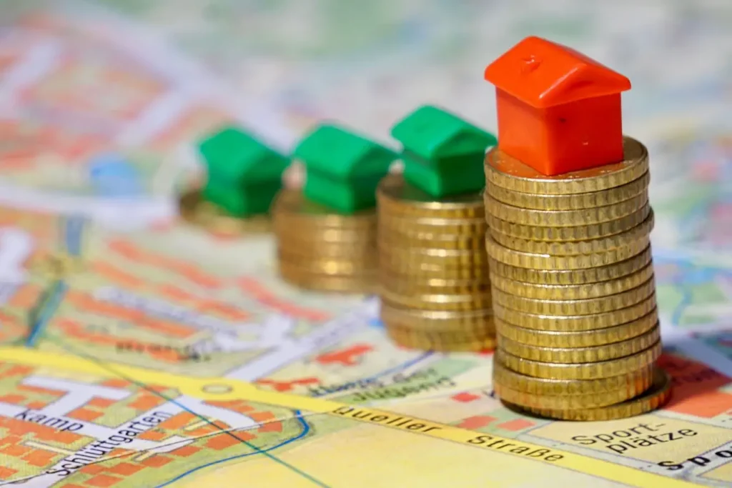 Petites maisons vertes et rouges posées sur des piles de pièces dorées, illustrant la montée des valeurs immobilières et l’effet psychologique des prix.