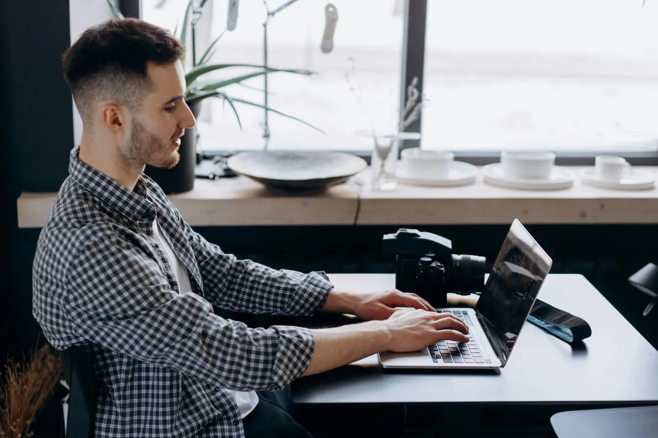 Personne en chemise à carreaux travaillant sur ordinateur avec appareil photo posé, image illustrant la recherche des premiers clients en freelance.