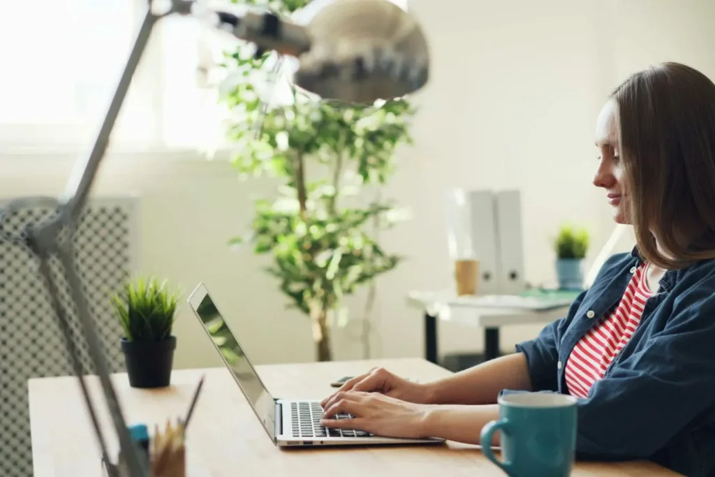 Personne tapant sur laptop posé sur bureau avec tasse bleue et plante, image illustrant l’usage d’outils de gestion de projet pour freelance.