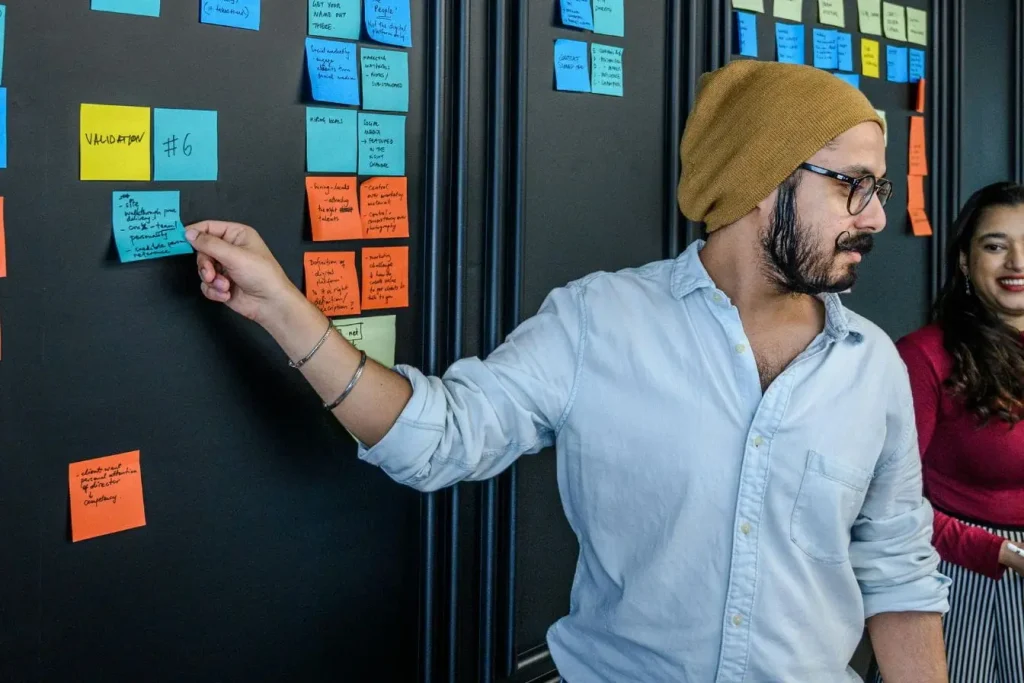 Personne en bonnet jaune plaçant post-it bleu sur mur sombre, image illustrant la méthode Design Thinking.