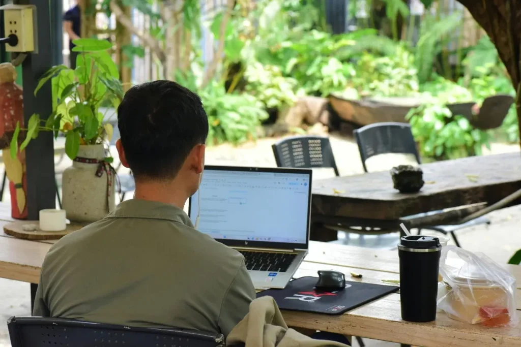Personne travaillant sur laptop en extérieur avec boisson et plantes, image illustrant la recherche des premiers clients en freelance dans un cadre inspirant.
