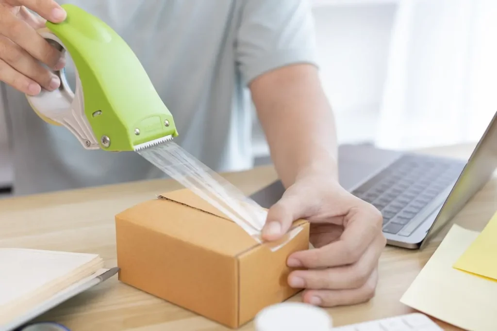 Entrepreneur prépare un colis sur bureau moderne avec laptop et carnet, illustrant la logistique rapide et essentielle du dropshipping.