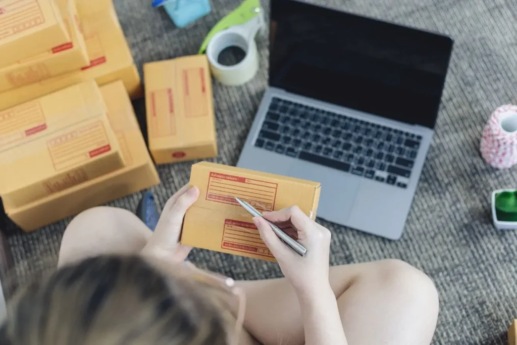 Personne assise entourée de cartons et laptop, écrit sur étiquette colis, image illustrant la préparation des commandes pour vendre sur Amazon.