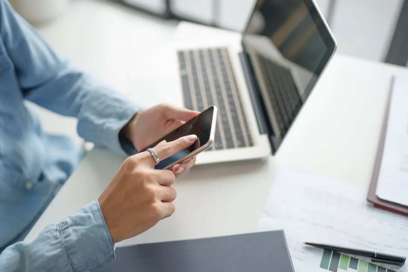 mobile-first indexing Personne assise devant un bureau utilisant un smartphone avec ordinateur ouvert, image qui illustre l’importance du mobile-first indexing dans le travail digital.